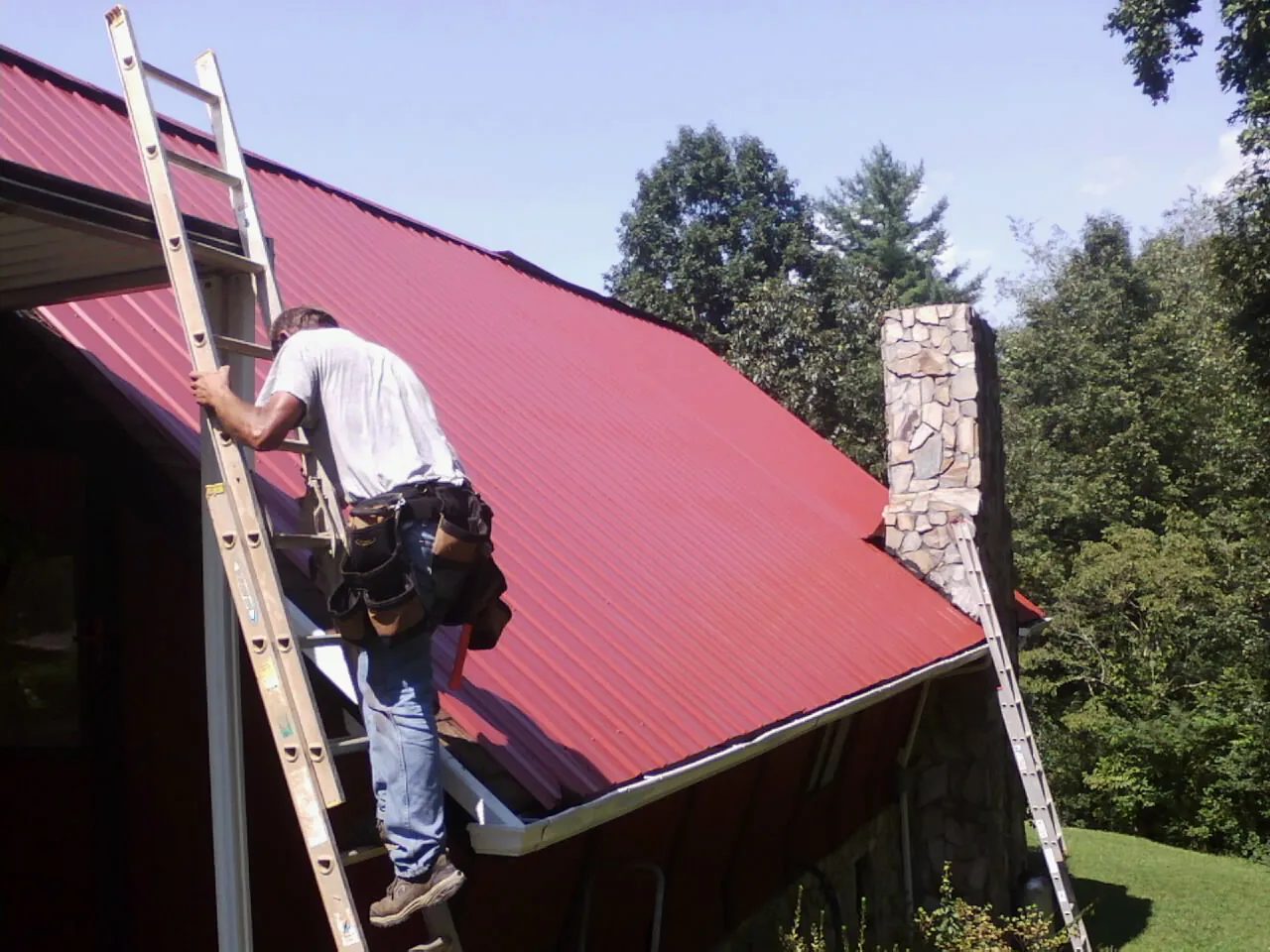 Roofer on a ladder beside a red metal roof and stone chimney — Soffit & Fascia Repair by Roofing Brothers in Rensselaerville